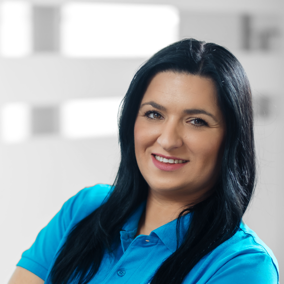 Smiling young woman with blonde hair and black glasses wearing a blue polo shirt against a blurred light background.